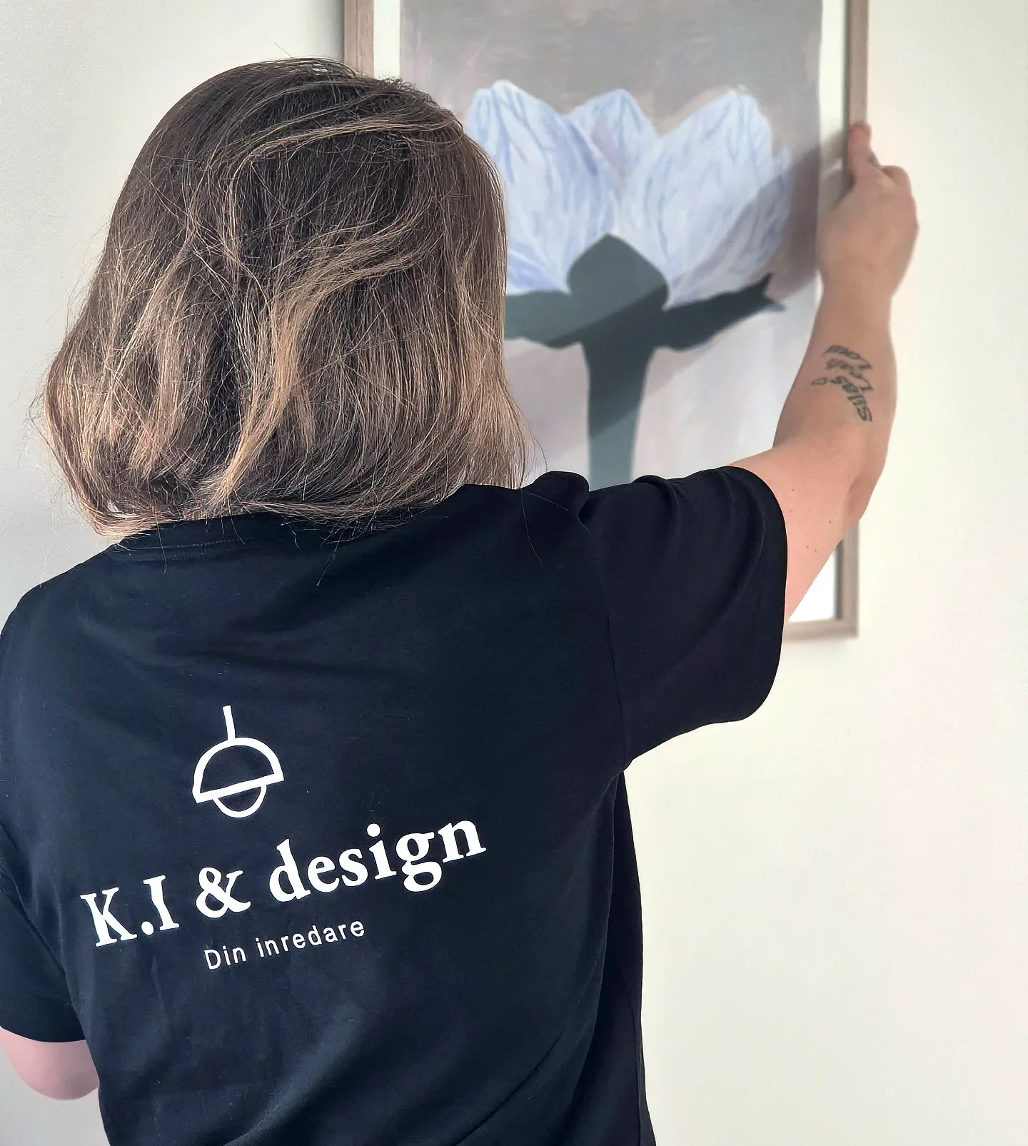 Person med K.I & design-tröja hänger upp en blomtavla på vägg.